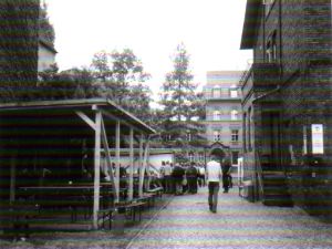 Dieses Foto zeigt links den Holzpavillion mit Sitzplätzen, rechts das Klostergebäude, wo sich die Kleiderkammer und Hygienestation befindet und die Sozialberatung stattfindet, mittig sieht man die Essensausgabe und die wartende Menschenschlange.